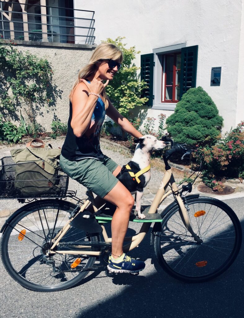 Frau sommerlich gekleidet fährt Fahrrad. Ihr Hund sitzt auf einem Brett zwischen ihren Beinen und fährt stolz mit.