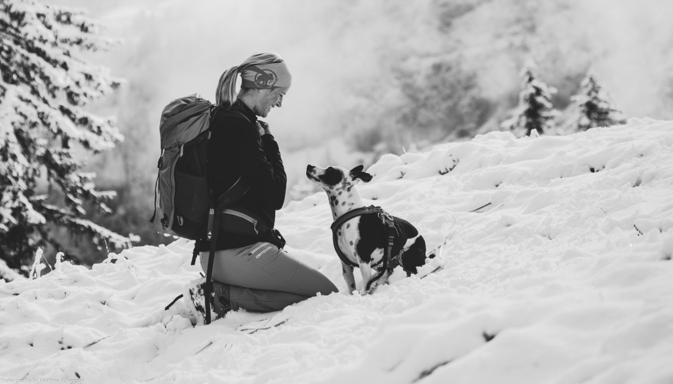 Frau sitzt frontal zu ihrem Hund in der Winterlandschaft. Hund blickt ihr direkt ins Gesicht. Frau lächelt glücklich.
